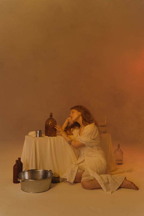 A Woman in White Dress Sitting on the Ground while Looking at the Glass Bottle Beside Her