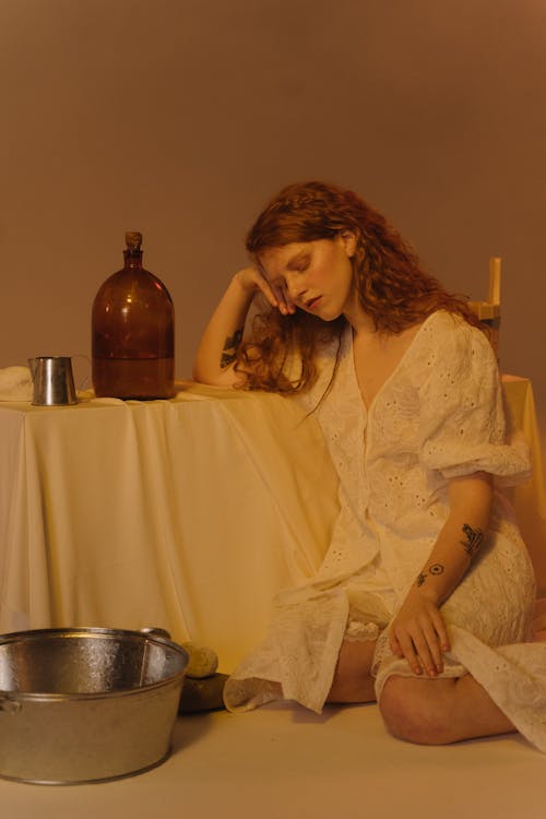 A Woman in a White Dress Sitting on the Floor while Leaning on a Table