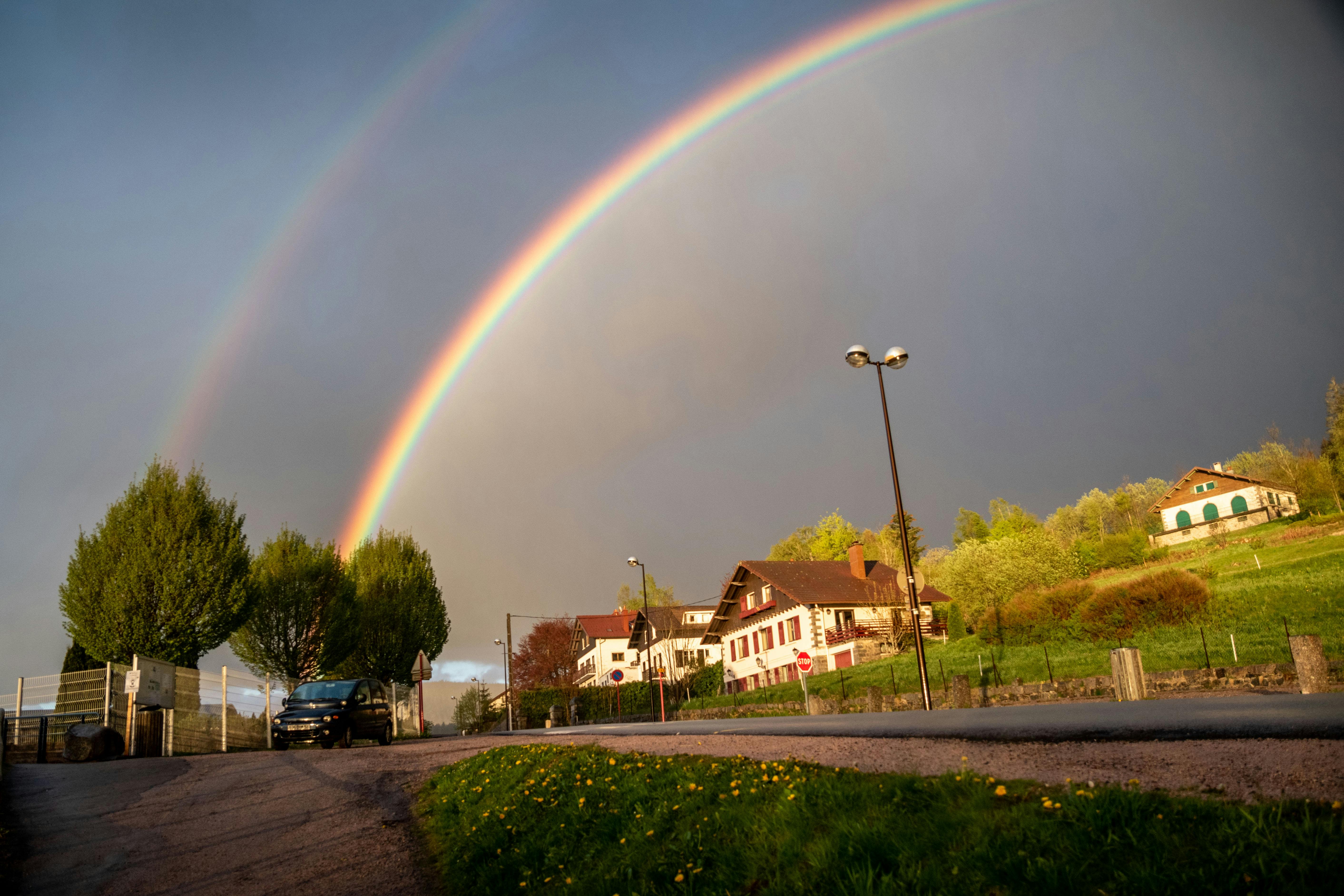 Vibrant double rainbow arches over charming countryside homes and a winding road.