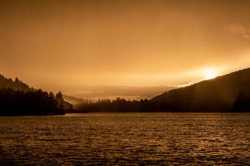 Free Scenic view of a golden sunset over a tranquil lake surrounded by mountains and forests. Stock Photo