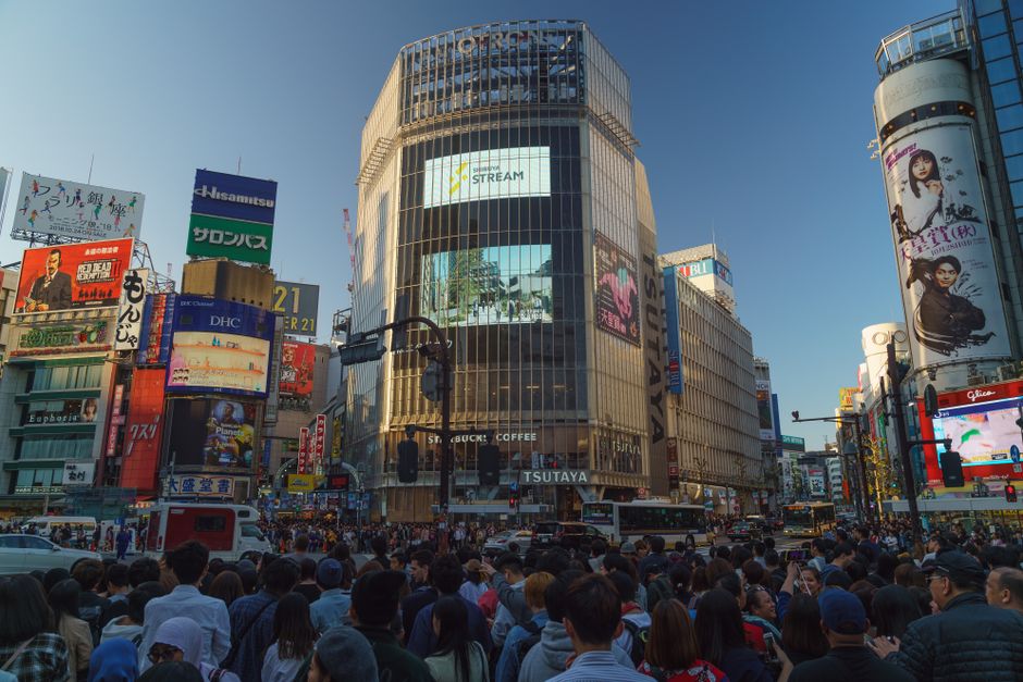 Crowded Shibuya Crossing in Tokyo captured during daytime with numerous billboards and bustling activity.