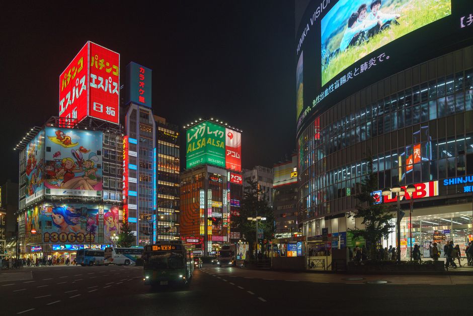 Lively street view of Shinjuku, Tokyo at night, featuring bright billboards and bustling city life.