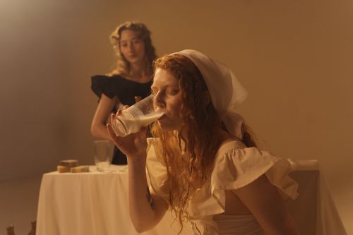Free A woman in a headscarf drinks milk indoors with another woman in the background, both in period costumes. Stock Photo