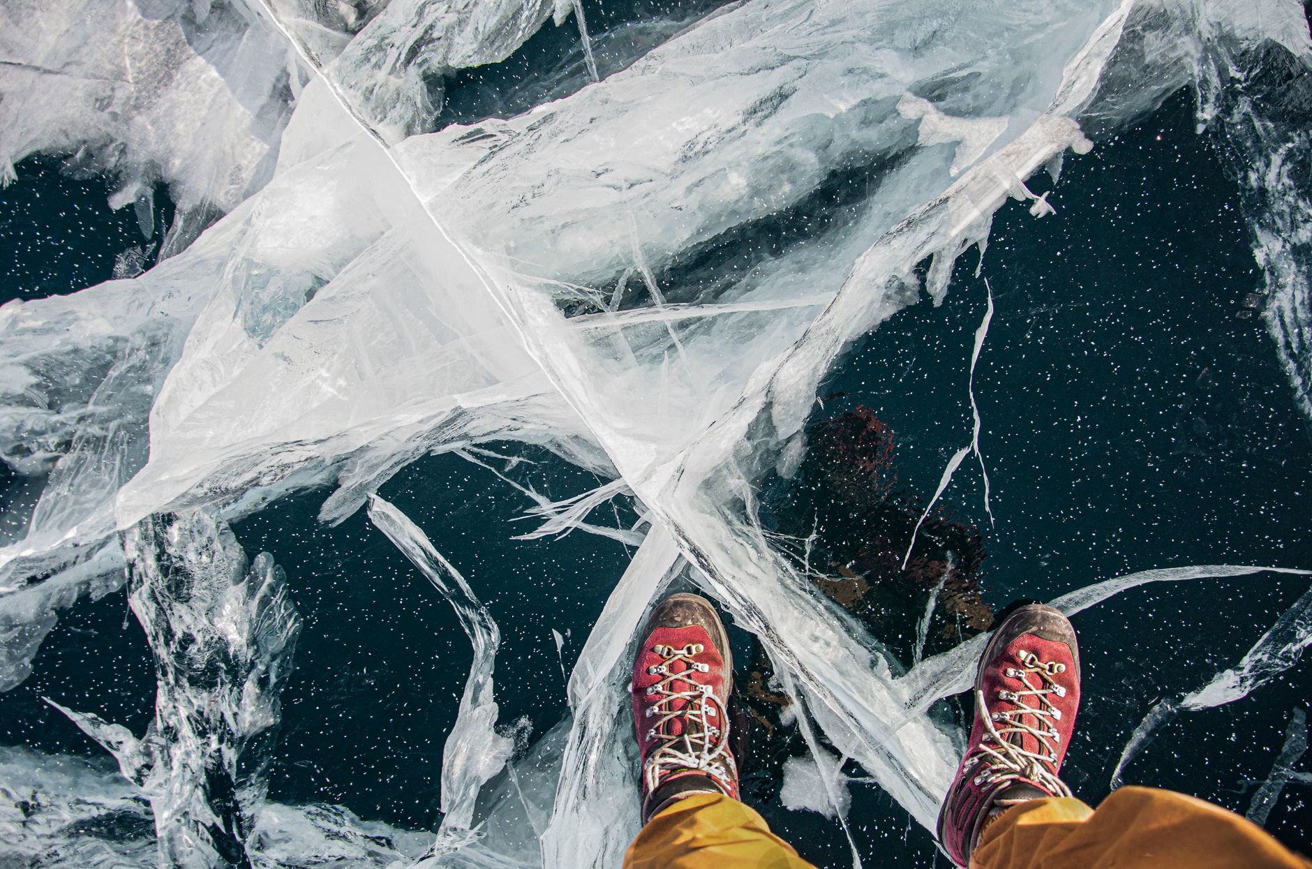A close-up view of red hiking boots standing on a cracked ice surface, showing intricate ice patterns.