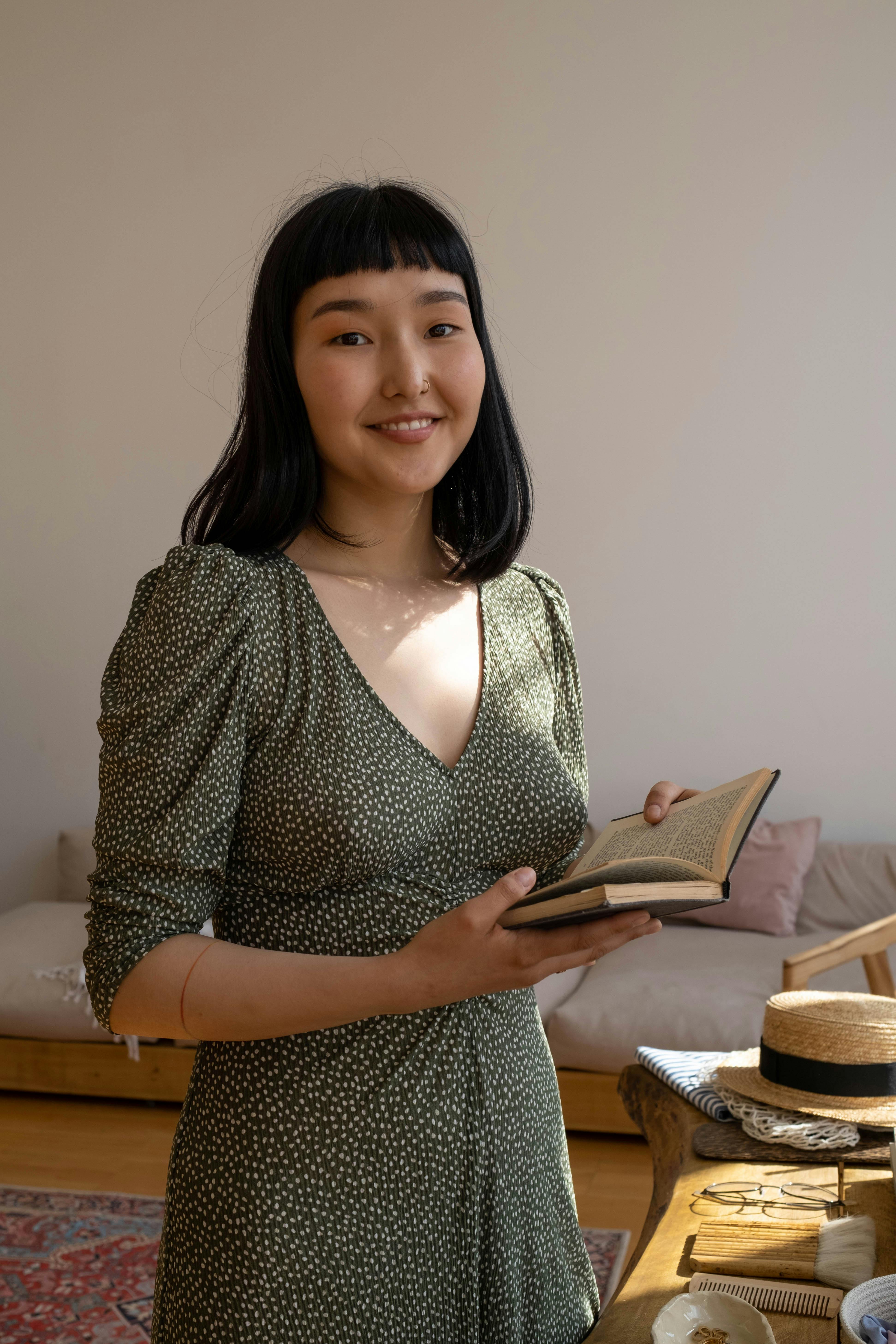 Asian woman in a green dress holding a book, smiling indoors with warm sunlight.