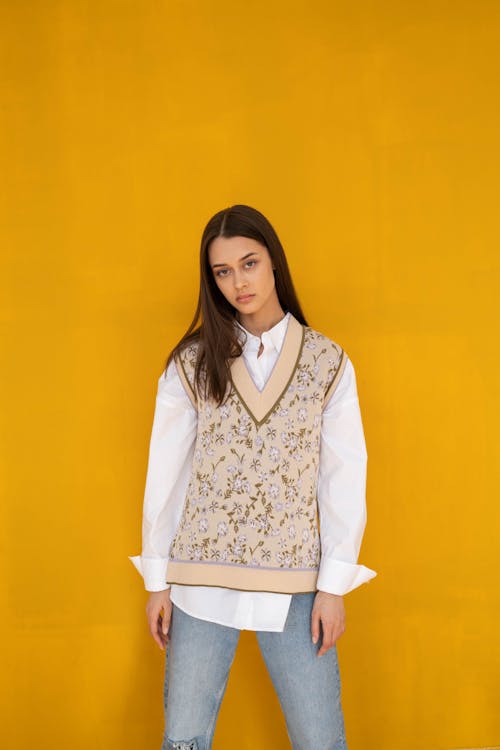 A Woman in White Long Sleeve Shirt Under a Floral Vest Standing Beside Yellow Wall