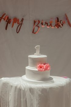 A minimalist white birthday cake with pink roses and a number one topper on a draped table.