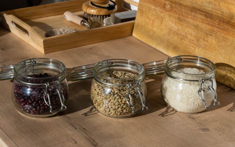 Glass jars of beans, lentils, and uncooked rice on a wooden table with warm lighting.