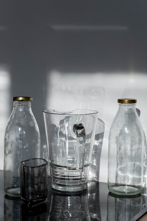 Free Clear Glass Pitcher Between Glass Bottles and Drinking Glass on Glass Surface Stock Photo