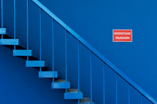 Blue staircase with safety sign reading 'Nooduitgang Vrijhouden.' Minimalist design.