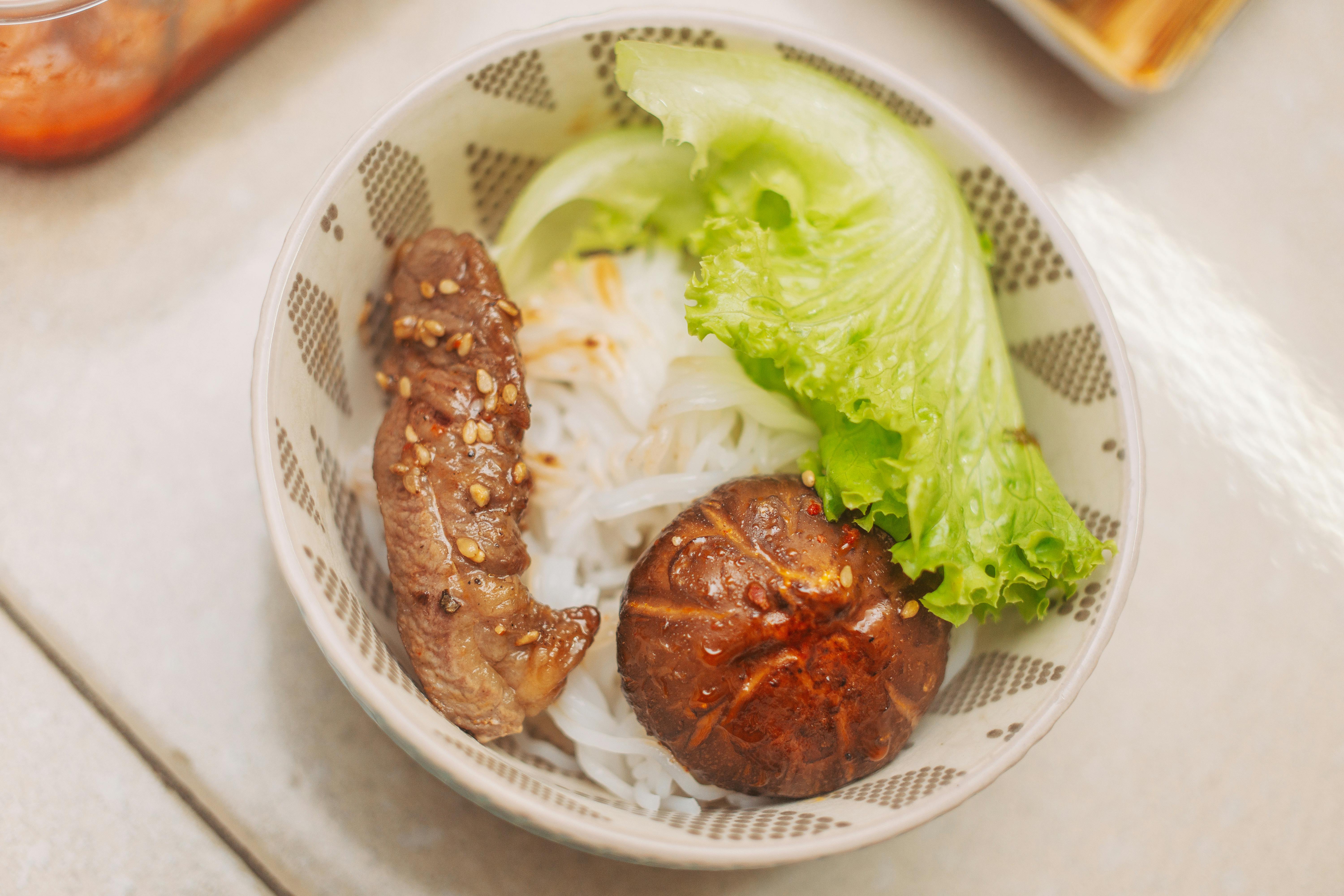 Delicious Vietnamese rice noodle bowl with steak, lettuce, and mushroom in a patterned bowl.