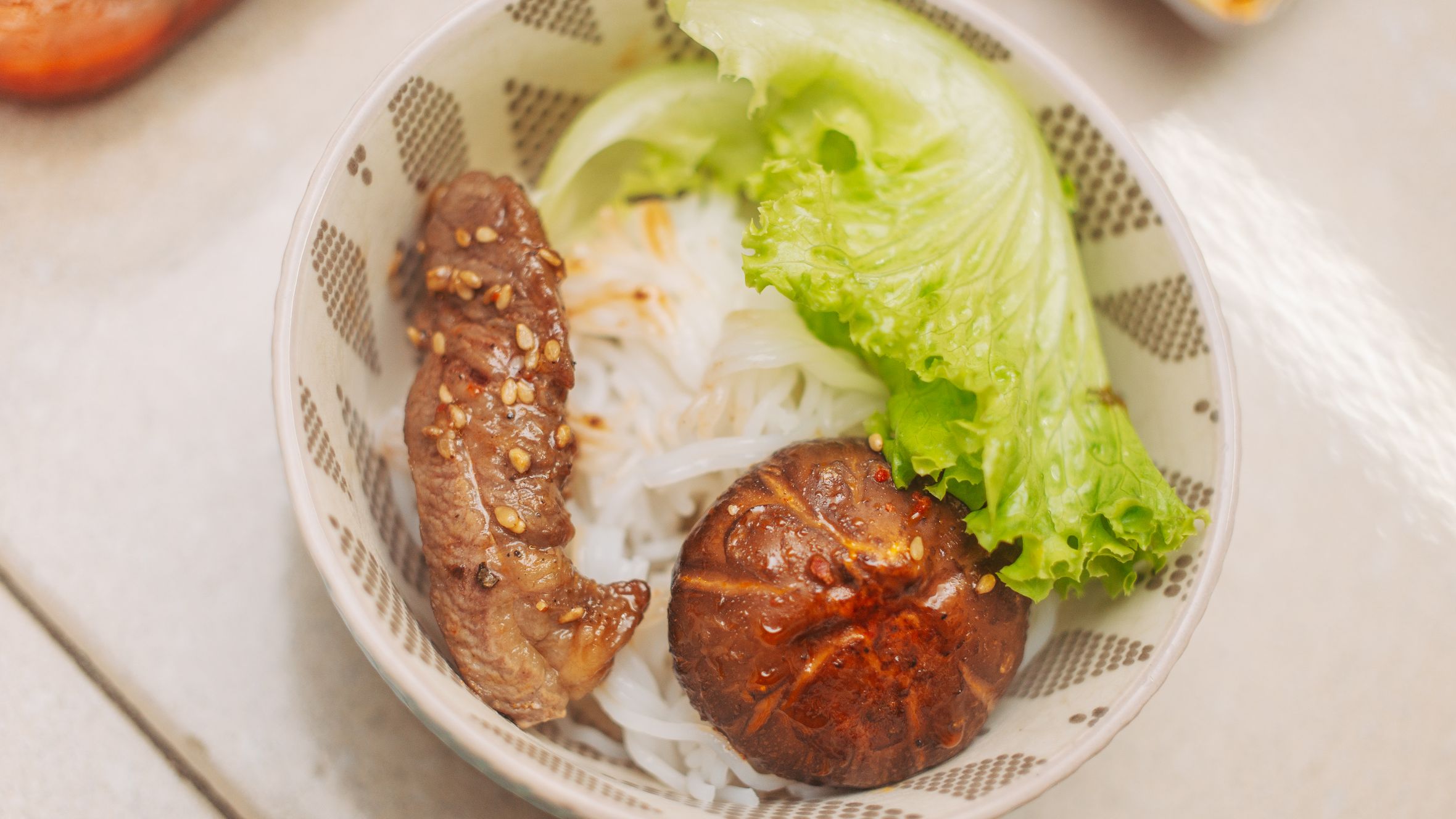 Delicious Vietnamese rice noodle bowl with steak, lettuce, and mushroom in a patterned bowl.