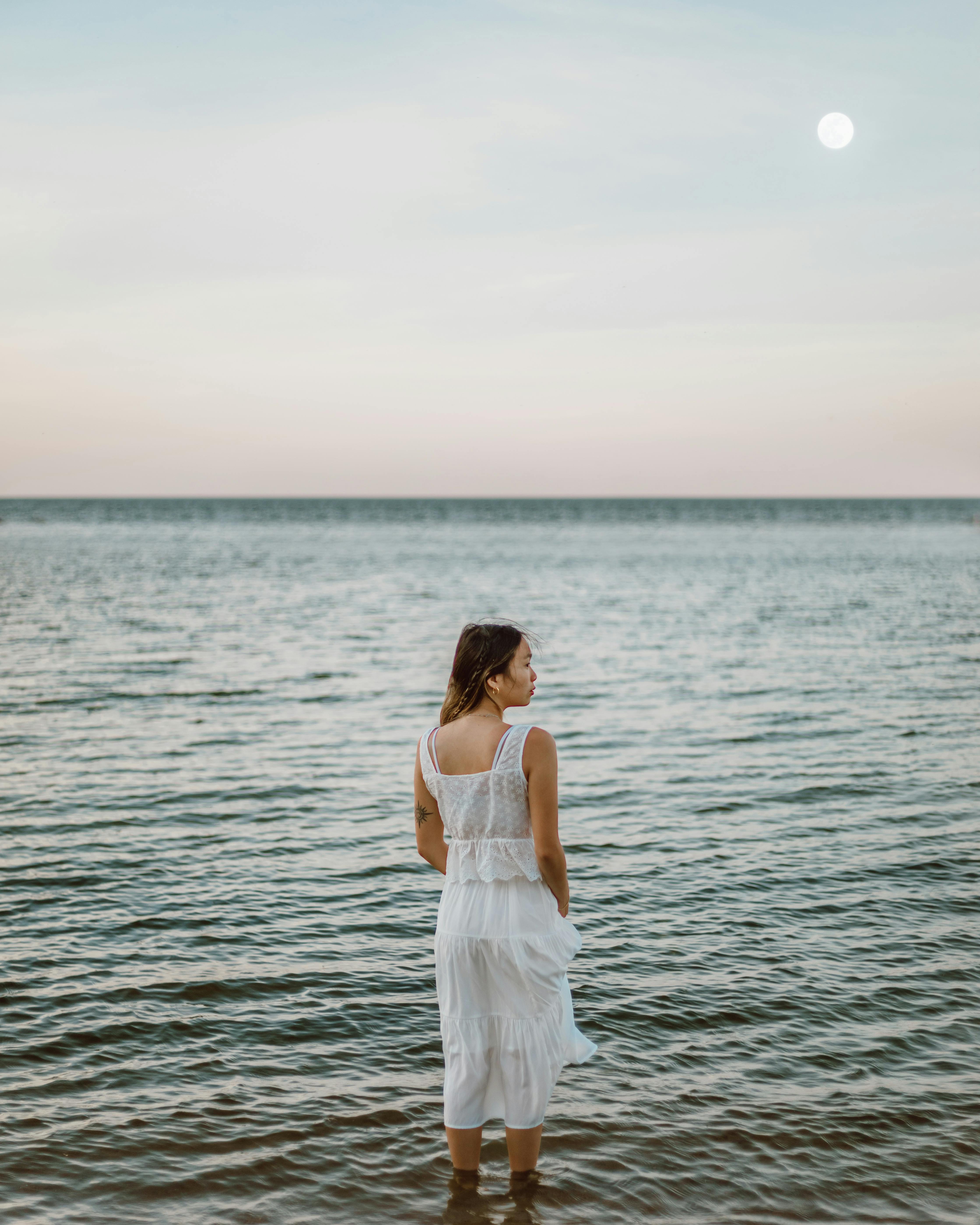 A woman in a white dress stands at the tranquil ocean during dusk.