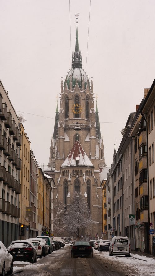 Free Snow-covered St. Paul's Church in Munich captured from a city street with parked cars and gothic architecture. Stock Photo
