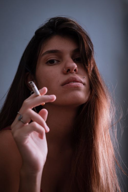 Free Close-up portrait of a young woman with piercing and cigarette, showcasing a moody expression. Stock Photo