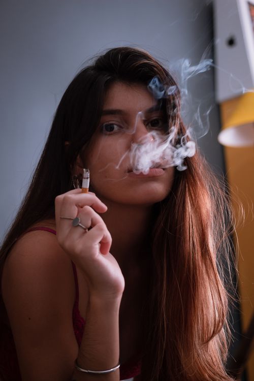 Free Young woman with long hair smoking indoors, exhaling smoke in a contemplative mood. Stock Photo
