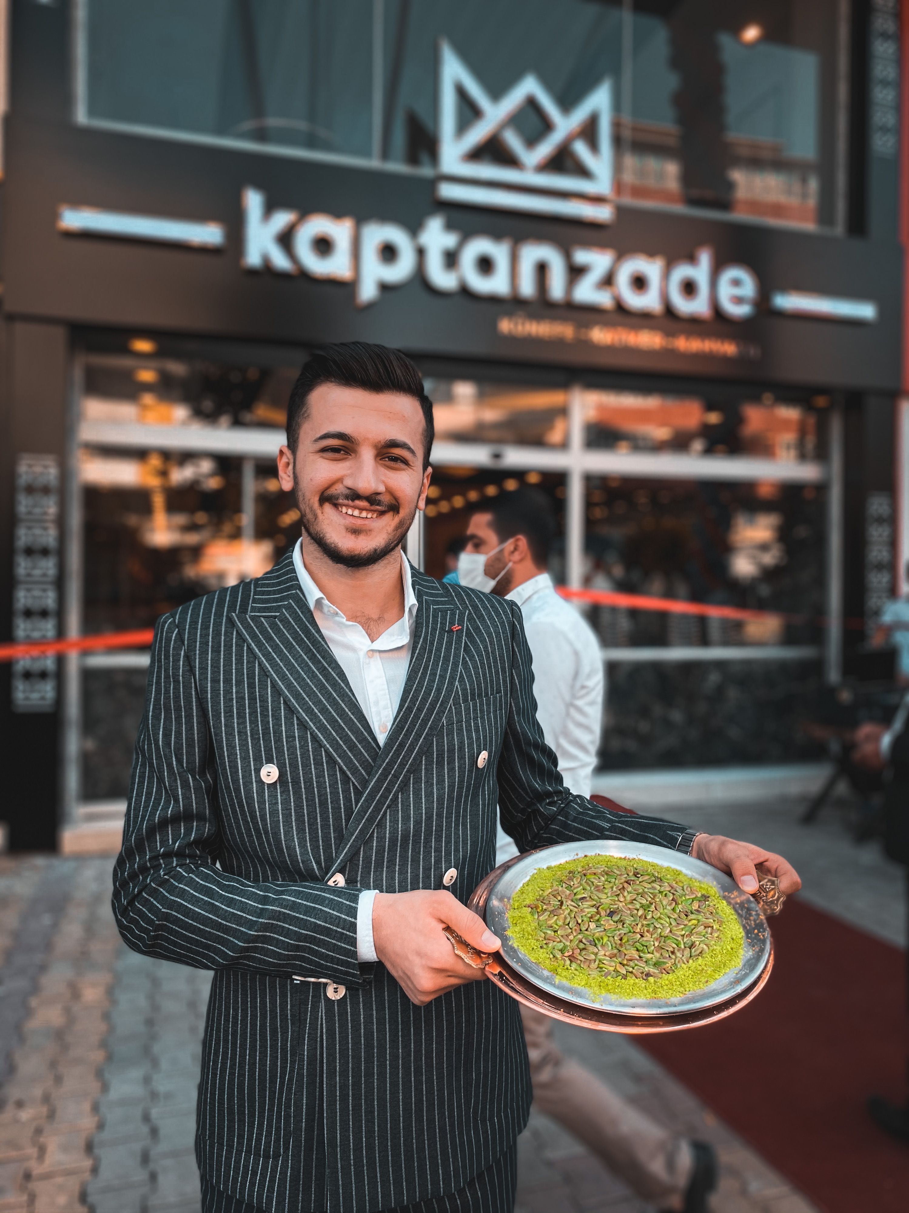 A smiling man in a striped suit holds a plate of Turkish dessert outside a restaurant.