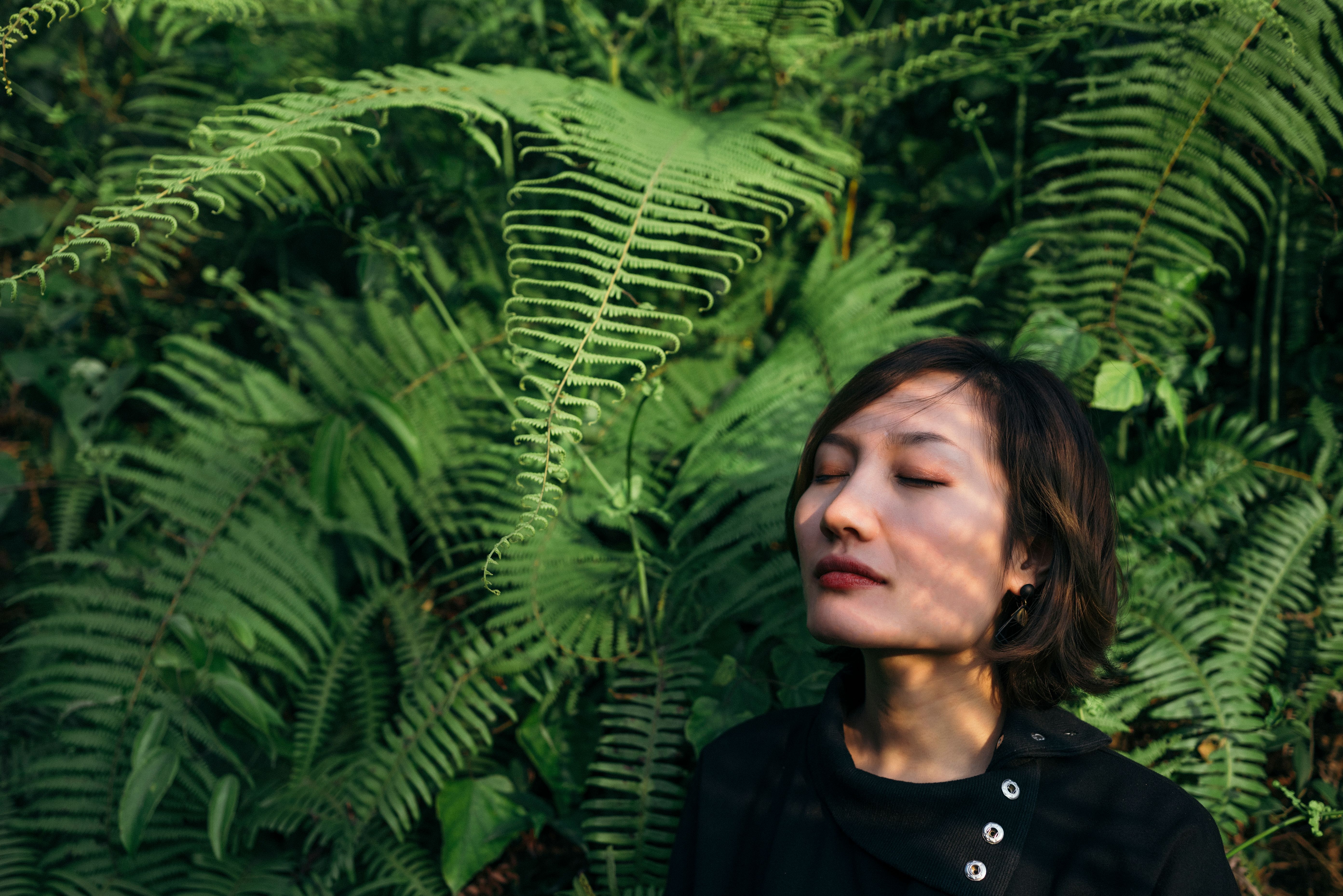 A tranquil scene of a woman with eyes closed, surrounded by lush fern leaves outdoors.