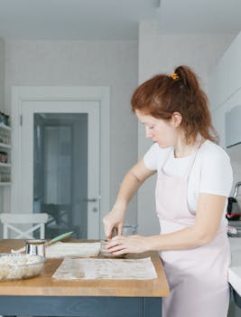 Woman in an apron making dough with a rolling pin at home kitchen.