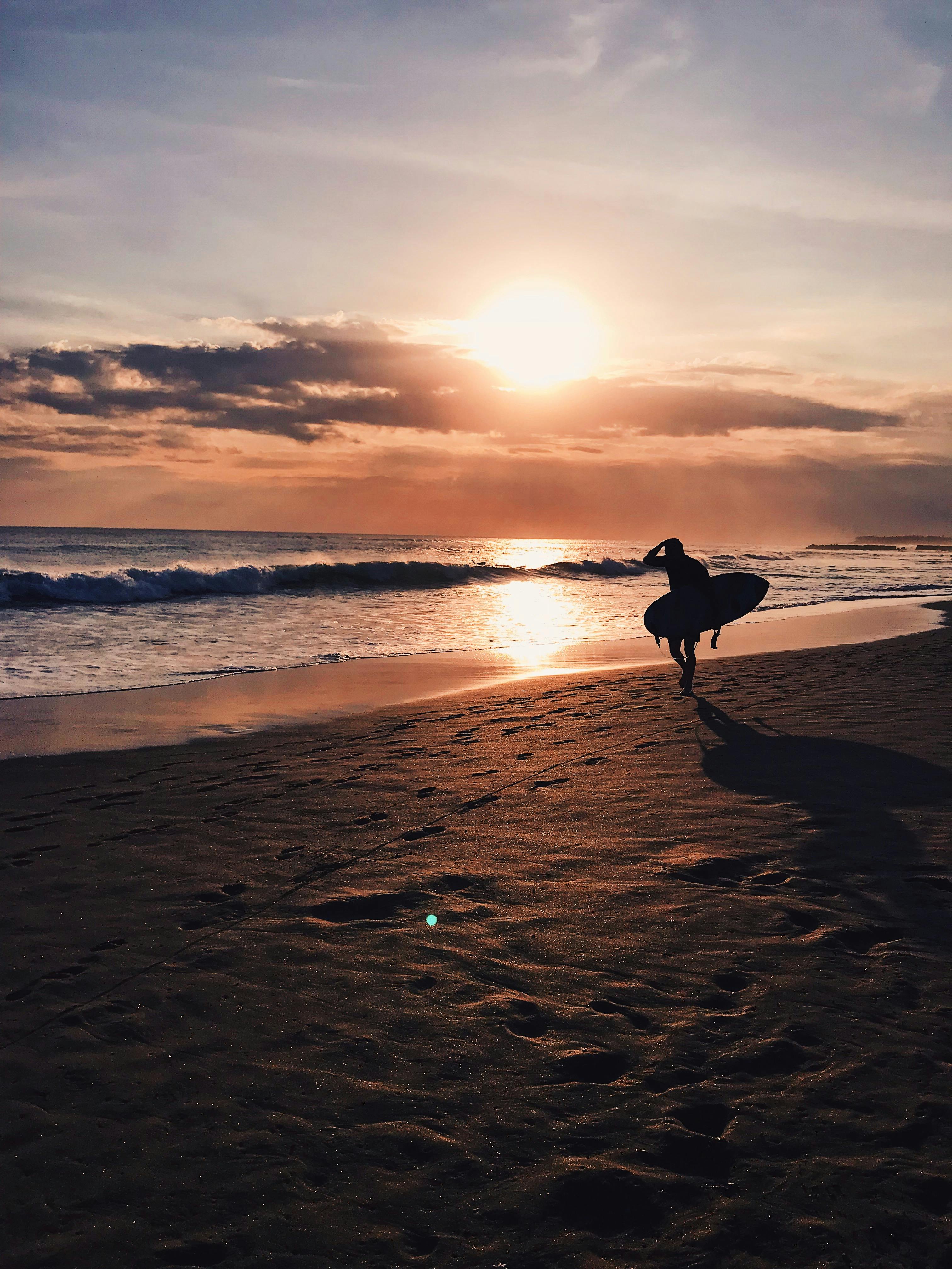 A silhouette of a surfer walking along the beach at sunset in Bali, Indonesia, capturing the golden hour's serene beauty.