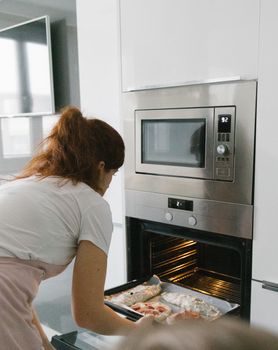 A woman placing a baking tray into an oven while cooking a homemade dish in a contemporary kitchen setting.