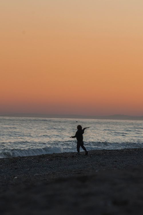 Free Silhouette of a child playing on a beach during a scenic sunset with an orange sky. Stock Photo
