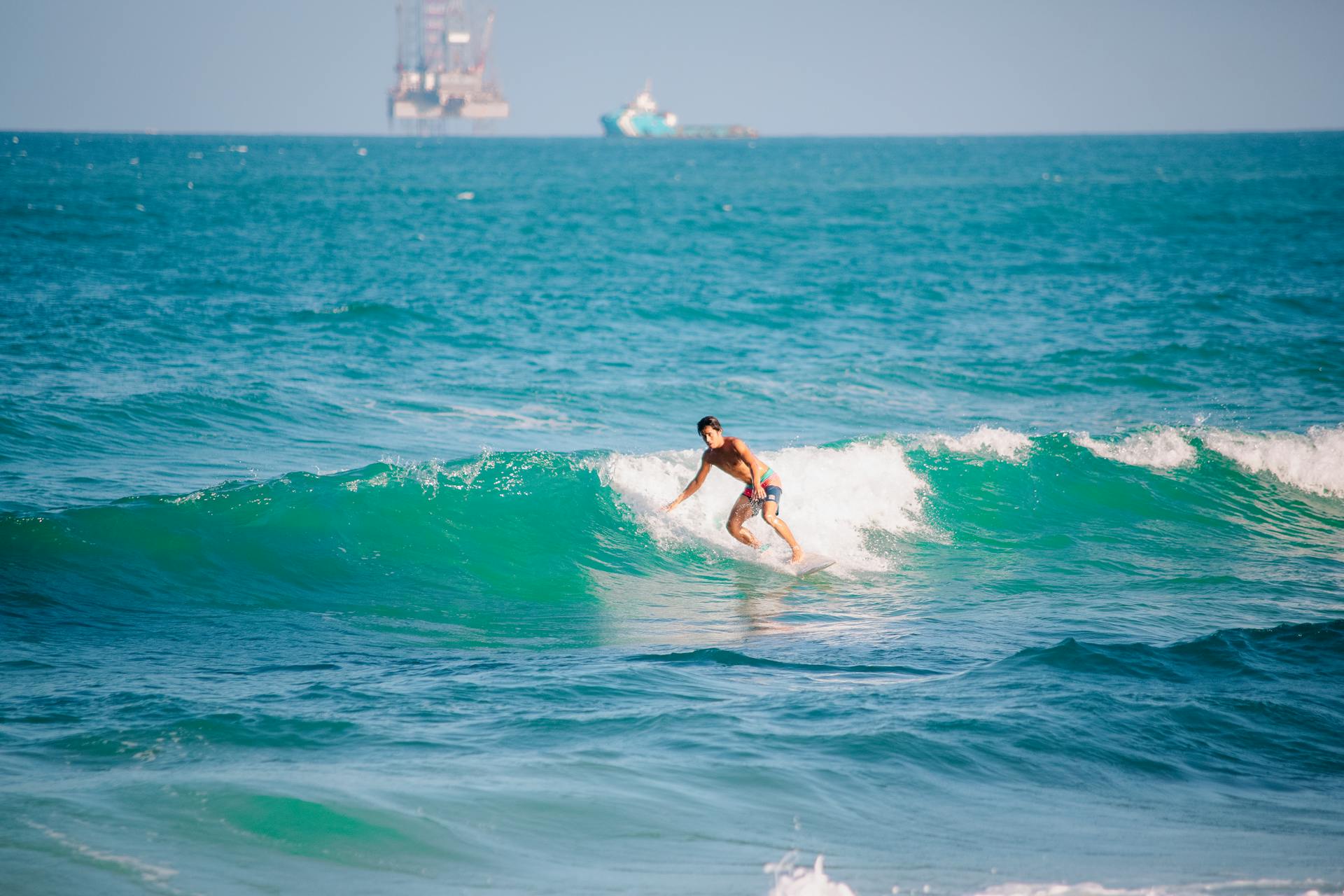 A surfer skillfully rides waves with an offshore platform in the background, capturing a sense of adventure and travel.