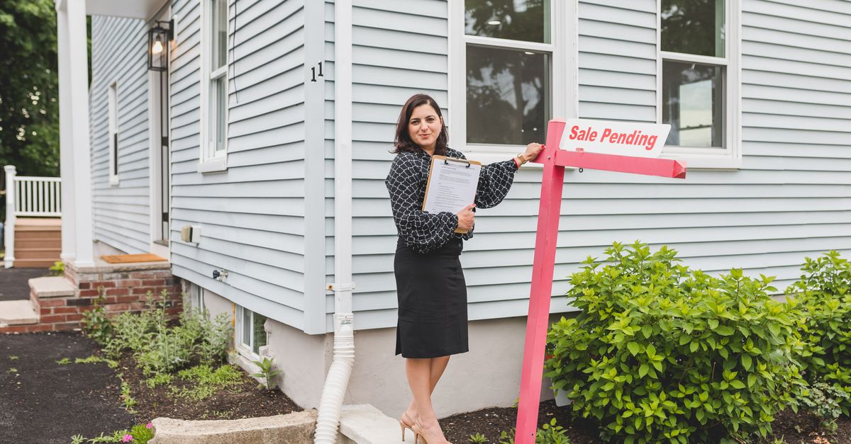 Real estate agent standing by a home with a sale pending sign, holding documents.
