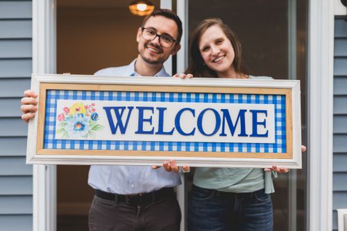 Free A smiling couple holds a cheerful welcome sign, expressing hospitality and warmth. Stock Photo