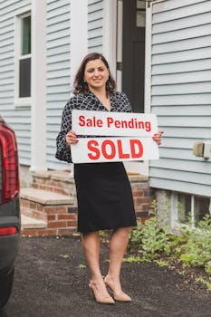Smiling real estate agent stands outside a house holding a 'Sold' sign, symbolizing property success.