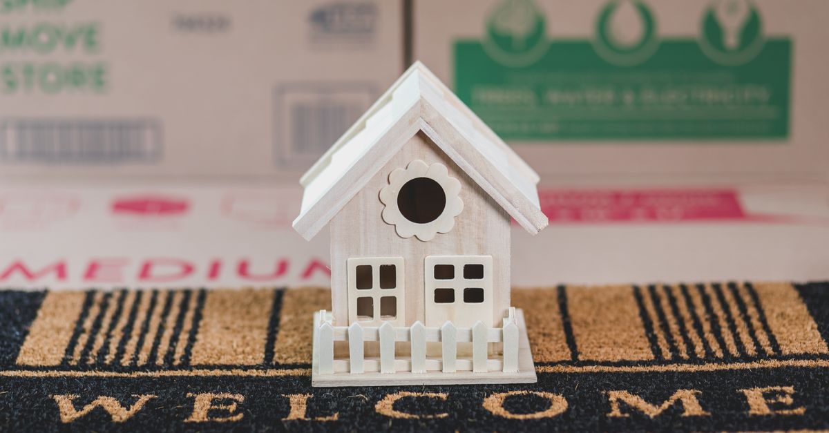 A small decorative wooden house on a welcome mat surrounded by moving boxes indoors.