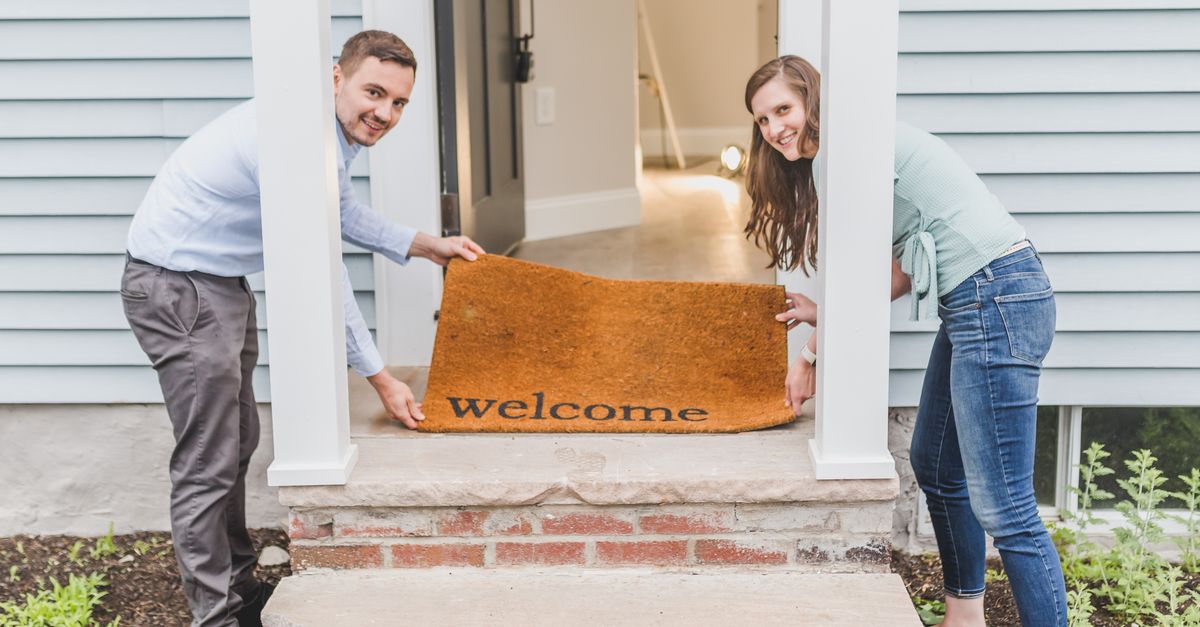 Couple smiling while holding a 'welcome' mat at their home's front door, symbolizing a warm greeting.