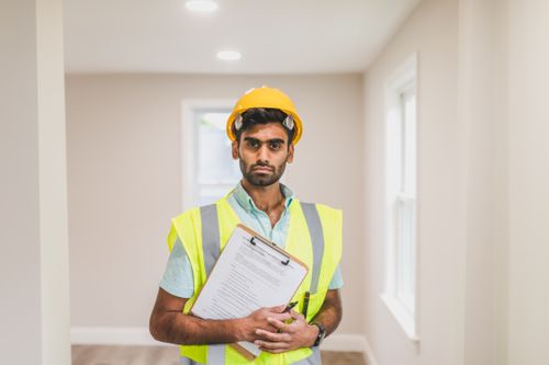 Free A construction worker wearing a hard hat and safety vest stands indoors with a clipboard. Stock Photo
