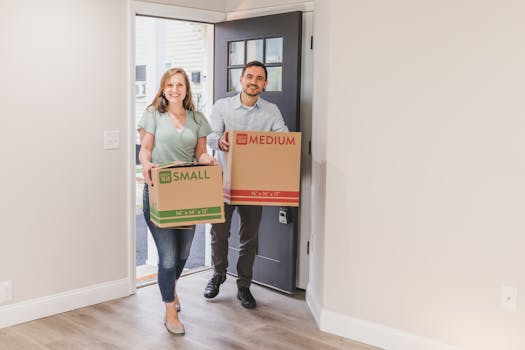 A smiling couple carrying cardboard boxes as they move into their new home.