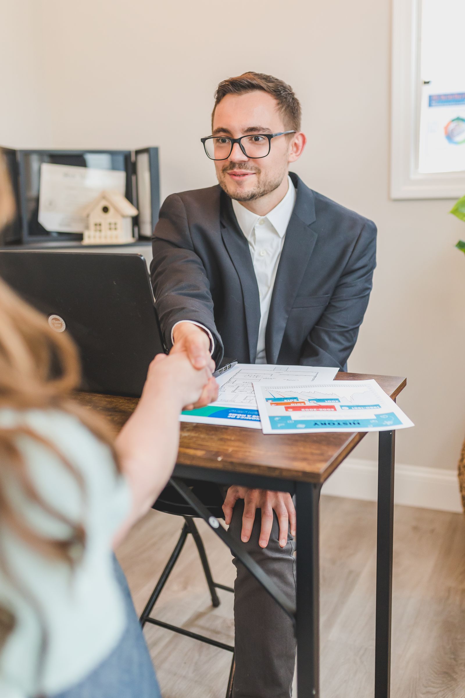 Free Real estate agent in suit shaking hands with client over property deal papers indoors. Stock Photo