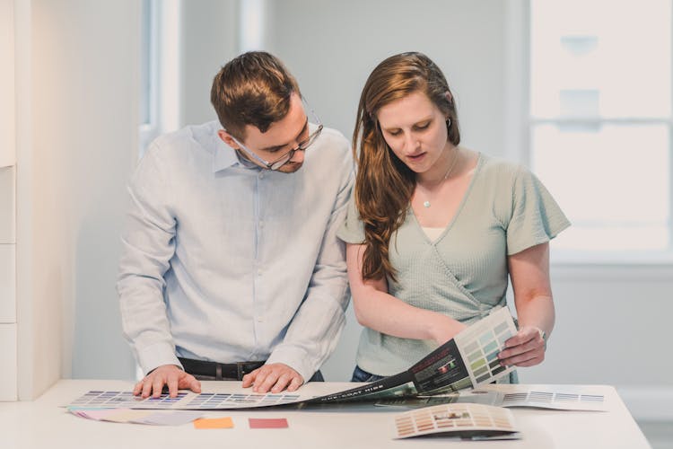 A Man And A Woman Standing Next To Each Other While Reading The Catalog