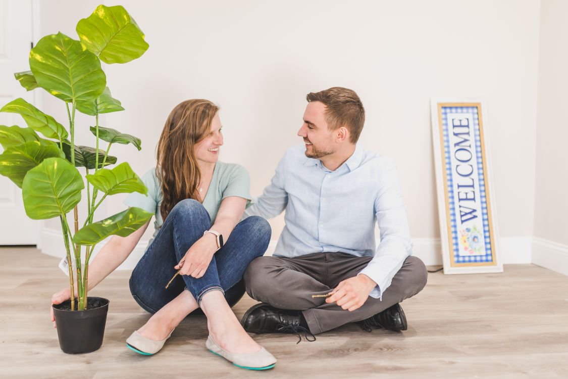Smiling Couple Sitting on Wooden Floor Beside a Houseplant and Welcome ...