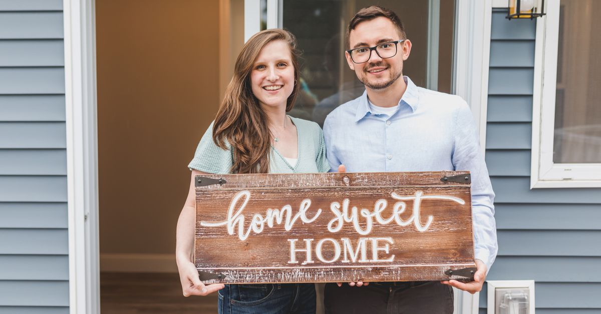 Smiling couple holding 'Home Sweet Home' sign in front of their new house entrance.