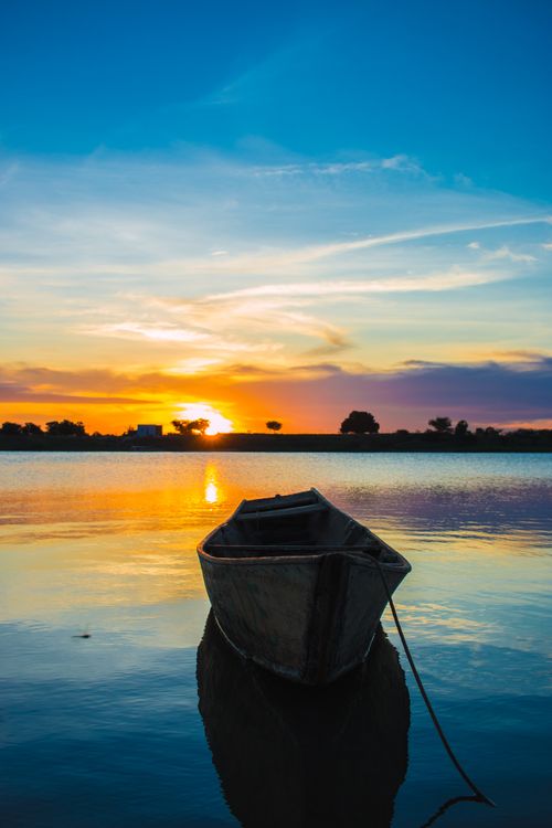 Free A calm sunset view over water featuring a solitary wooden boat reflecting in the evening light. Stock Photo