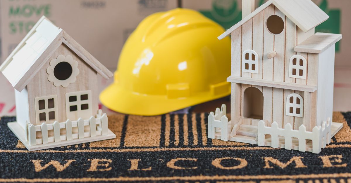 Miniature wooden houses on a welcome mat with a yellow hard hat in the background, symbolic of home building or construction.