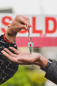 Close-up of hands exchanging house key with colorful SOLD sign in the background.
