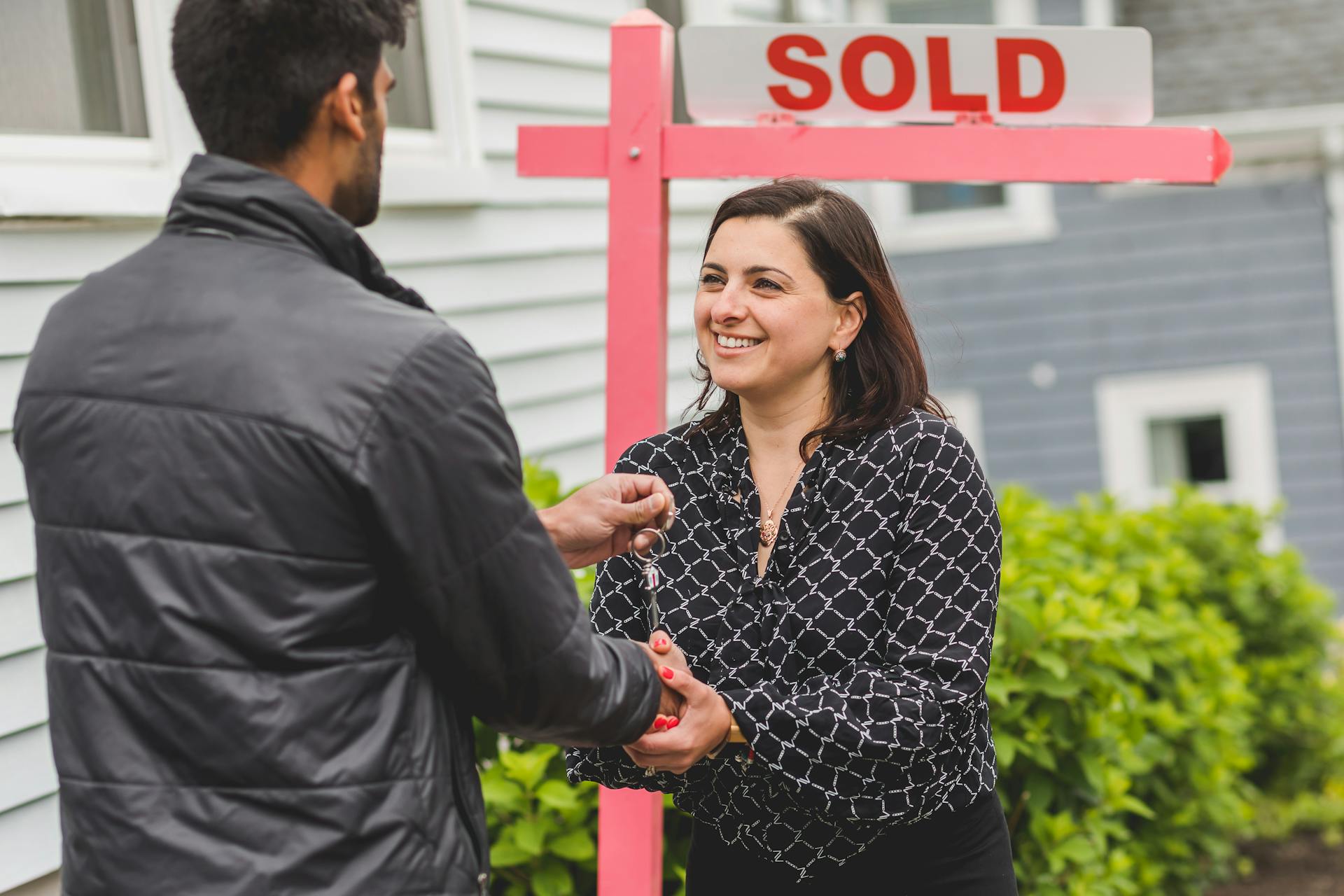 Real estate agent handing keys to a new homeowner with a 'Sold' sign in the background.