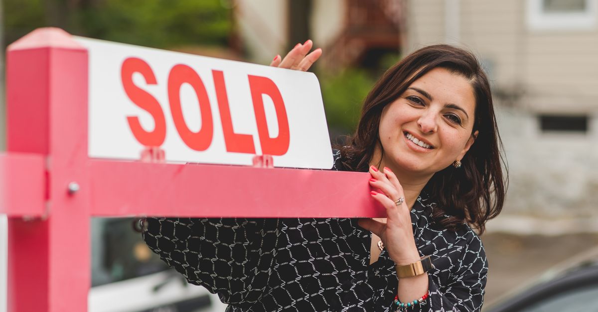 Smiling realtor holding a 'Sold' sign in front of a house. Perfect for real estate marketing.