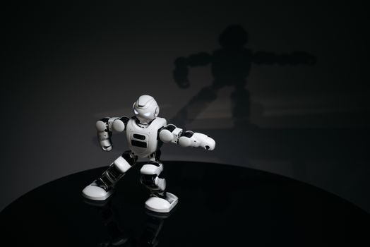 A futuristic white toy robot striking a pose in a dimly lit studio with a shadow cast behind it.