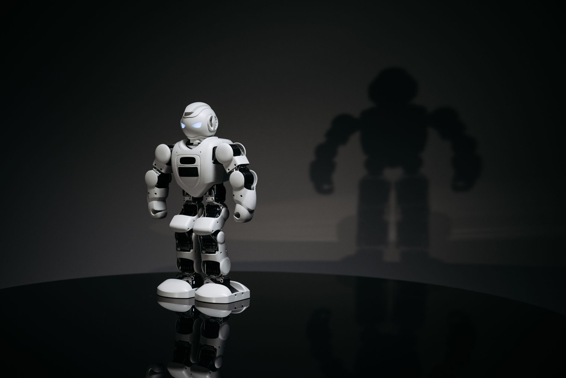 A white and black toy humanoid robot in a studio setting casting a shadow.