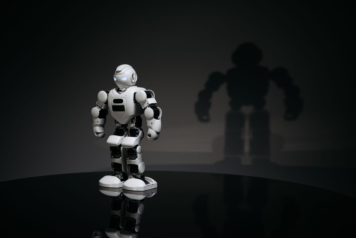 A white and black toy humanoid robot in a studio setting casting a shadow.