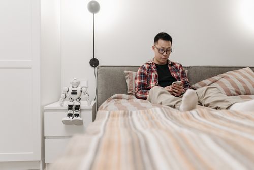 Free A man sits on a bed using a smartphone next to a robot toy, indoors. Stock Photo