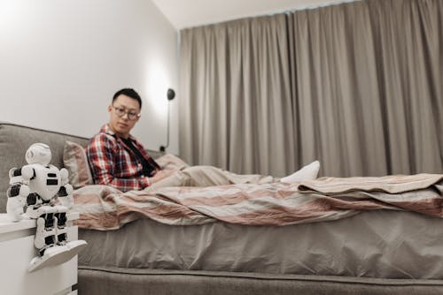 A Man Sitting on His Bed while Looking at the Robot