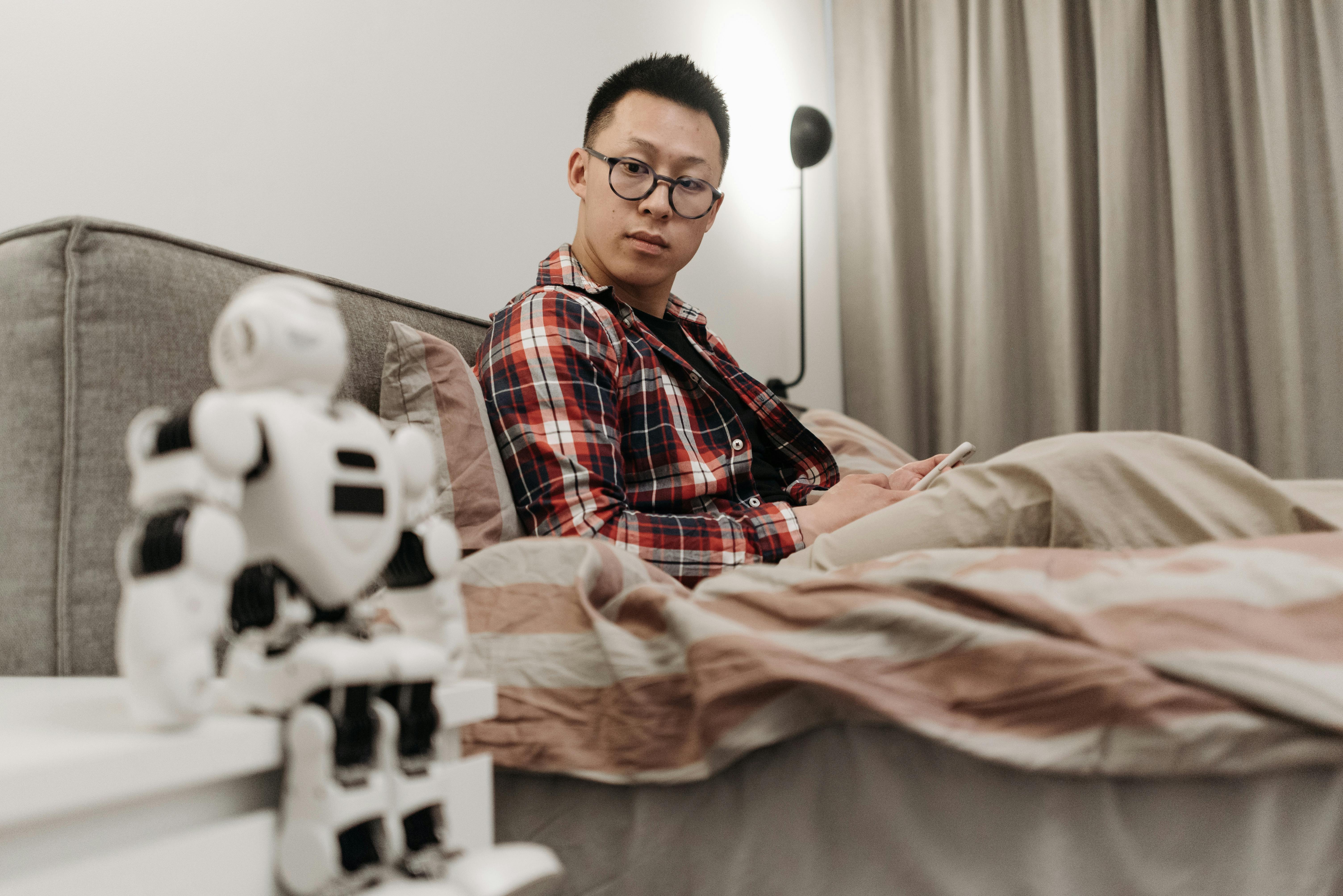 man with eyeglasses sitting on bed looking at a toy robot