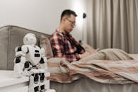 A Man Sitting on the Bed Near the White Robot on the Bedside Table